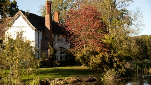 Timber-framed manor house with moat in foreground and red-leaved tree to one side